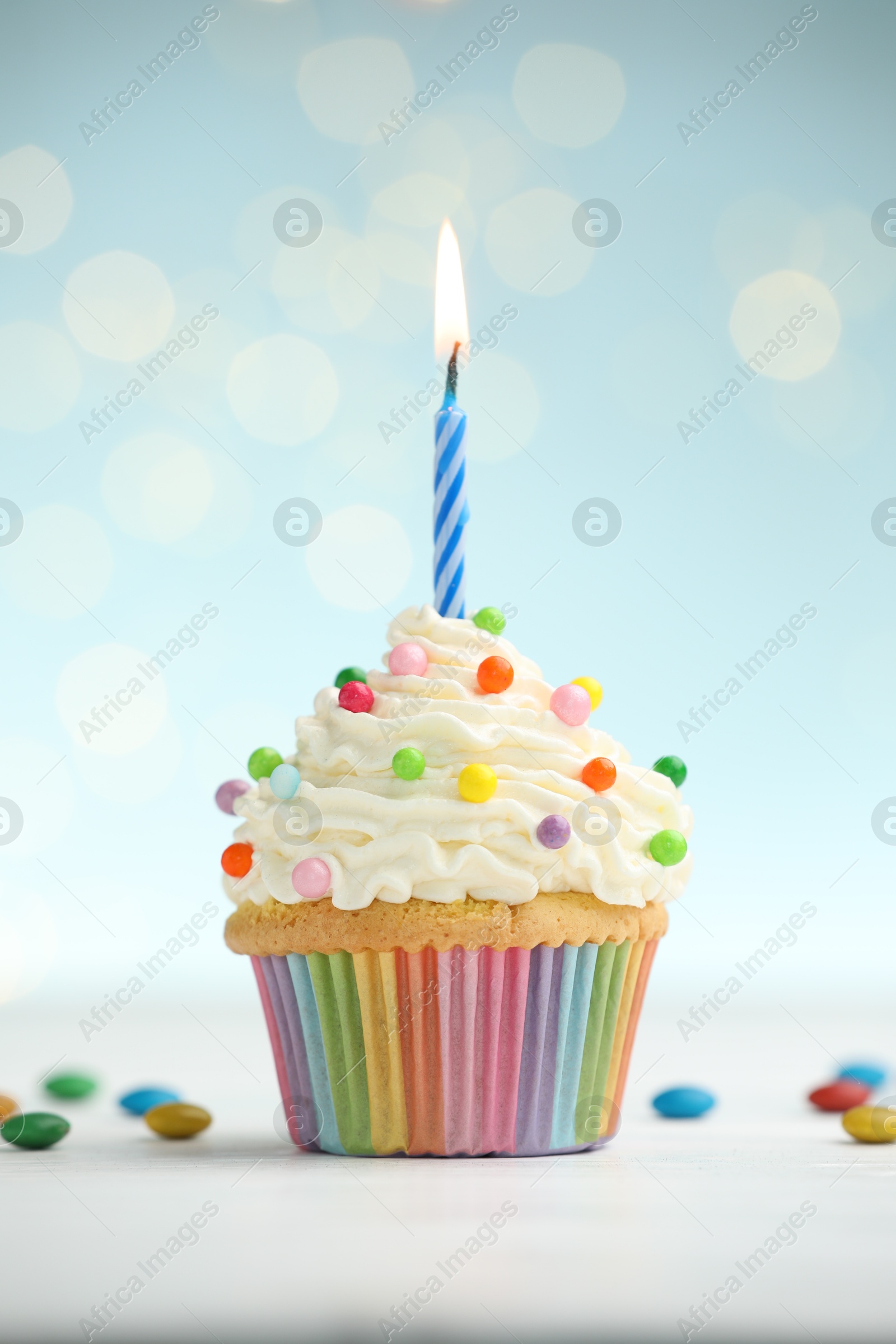 Photo of Burning candle in cupcake and candies on table against blue background with blurred lights