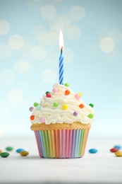Photo of Burning candle in cupcake and candies on table against blue background with blurred lights