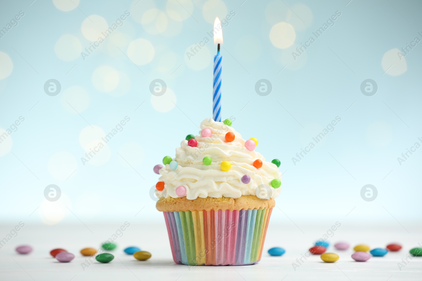 Photo of Burning candle in cupcake and candies on table against blue background with blurred lights