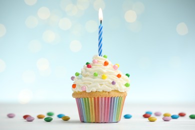 Photo of Burning candle in cupcake and candies on table against blue background with blurred lights