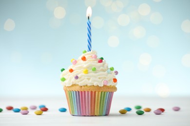 Photo of Burning candle in cupcake and candies on table against blue background with blurred lights