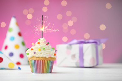 Photo of Burning sparkler in cupcake and party decor on table against pink background with blurred lights. Space for text