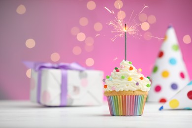 Photo of Burning sparkler in cupcake and party decor on table against pink background with blurred lights. Space for text