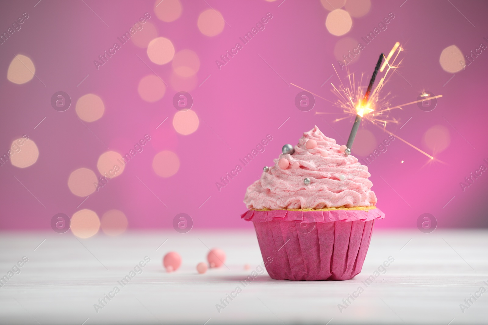 Photo of Burning sparkler in cupcake on table against pink background with blurred lights. Space for text