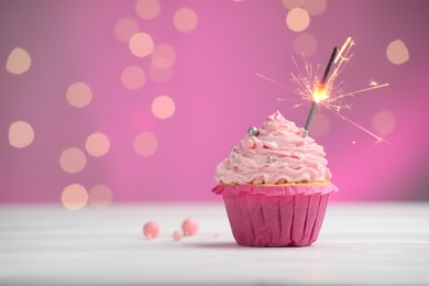 Photo of Burning sparkler in cupcake on table against pink background with blurred lights. Space for text