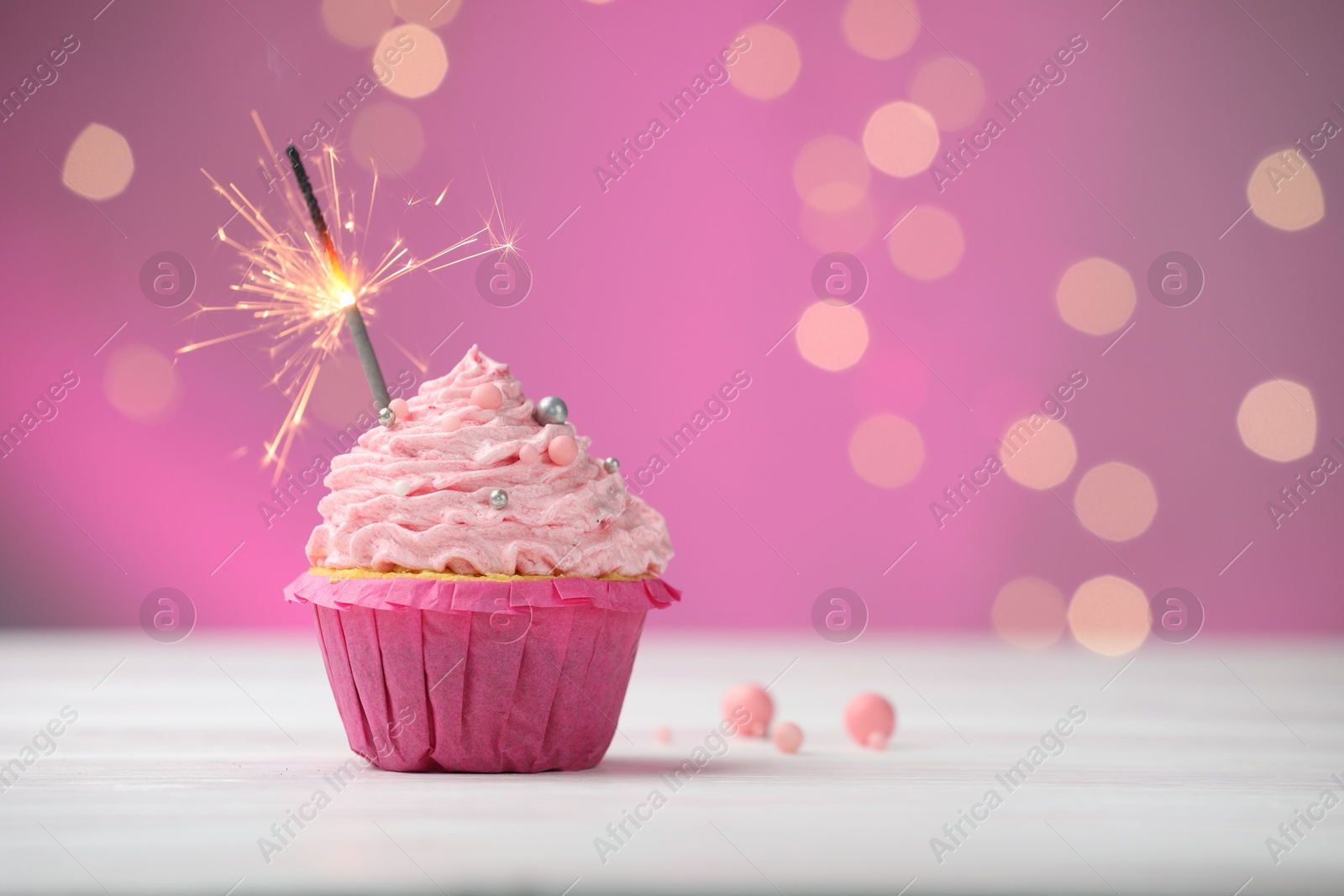 Burning sparkler in cupcake on table against pink background with blurred lights. Space for text Photo of Burning sparkler in cupcake on table against pink background with blurred lights. Space for text