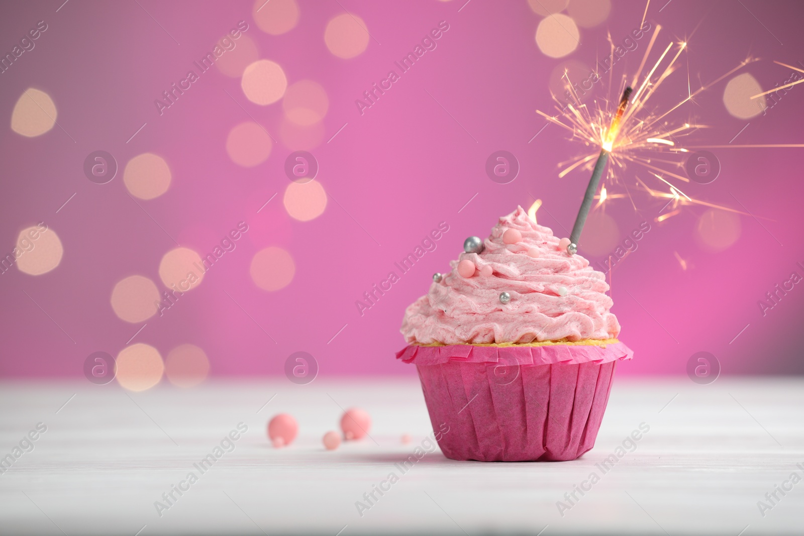 Photo of Burning sparkler in cupcake on table against pink background with blurred lights. Space for text