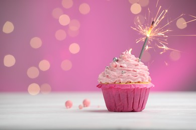 Photo of Burning sparkler in cupcake on table against pink background with blurred lights. Space for text
