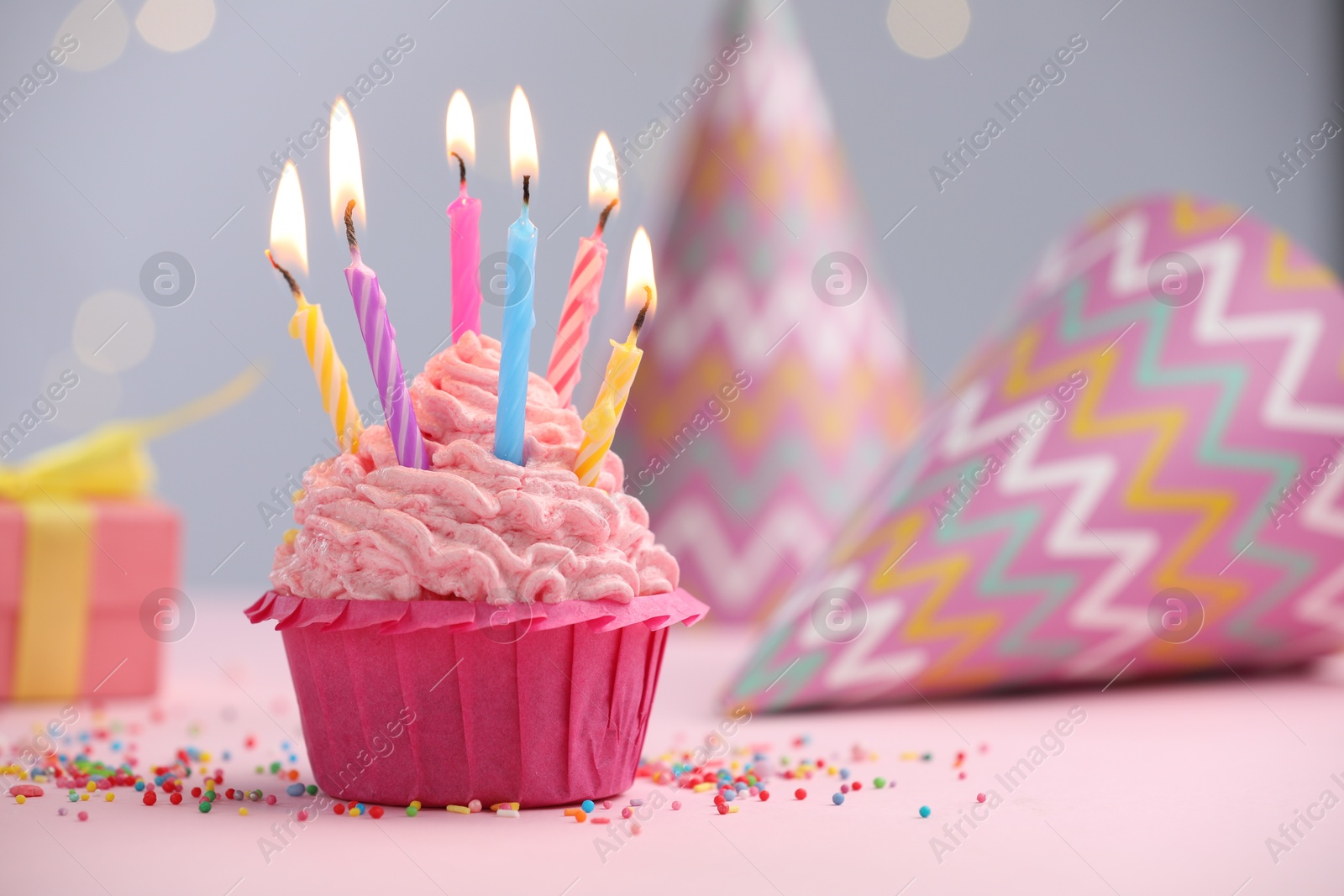 Photo of Burning candles in cupcake, sprinkles and party decor on table against blurred lights