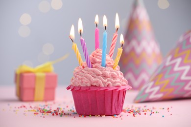 Photo of Burning candles in cupcake, sprinkles and party decor on table against blurred lights