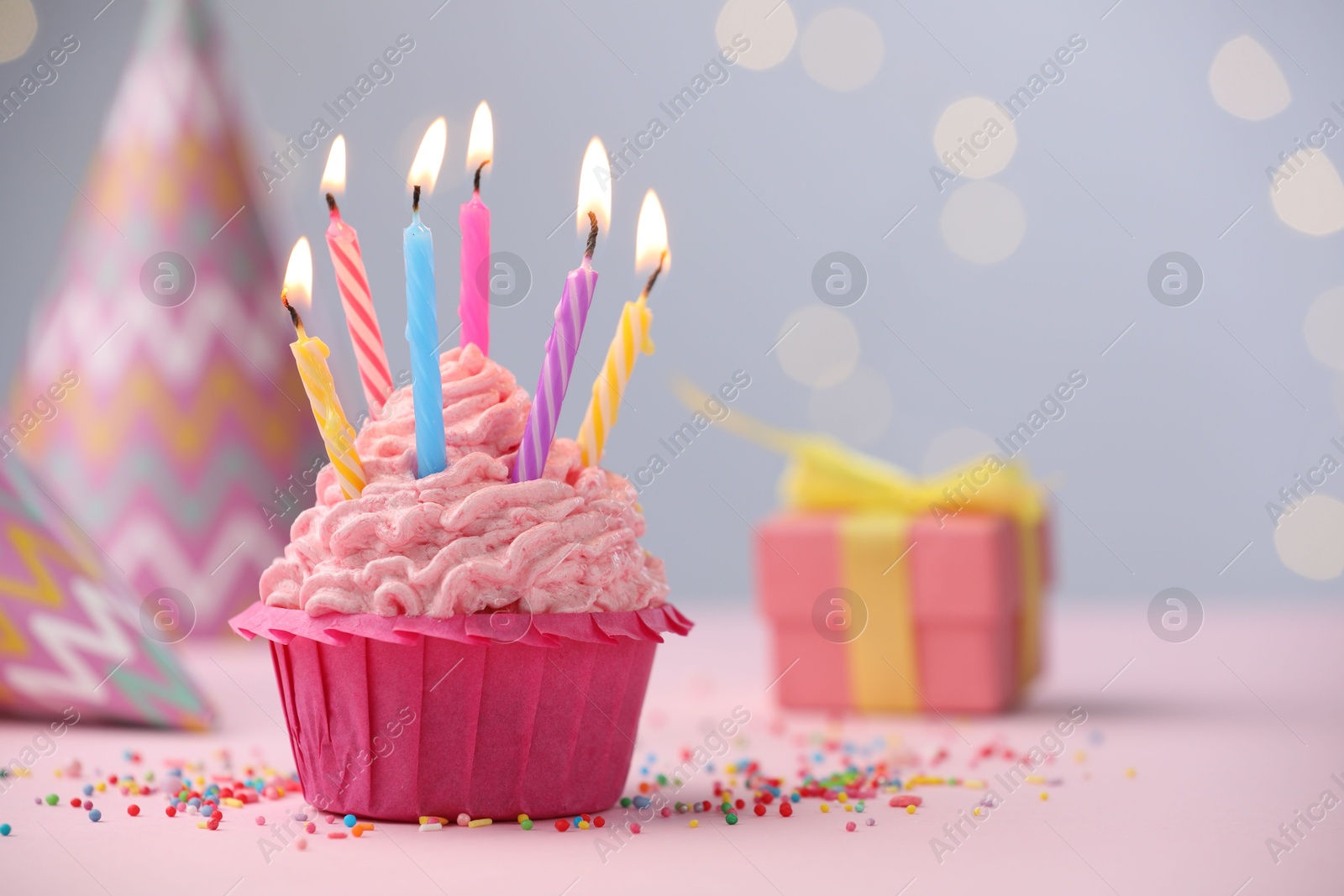 Photo of Burning candles in cupcake, sprinkles and party decor on table against blurred lights. Space for text