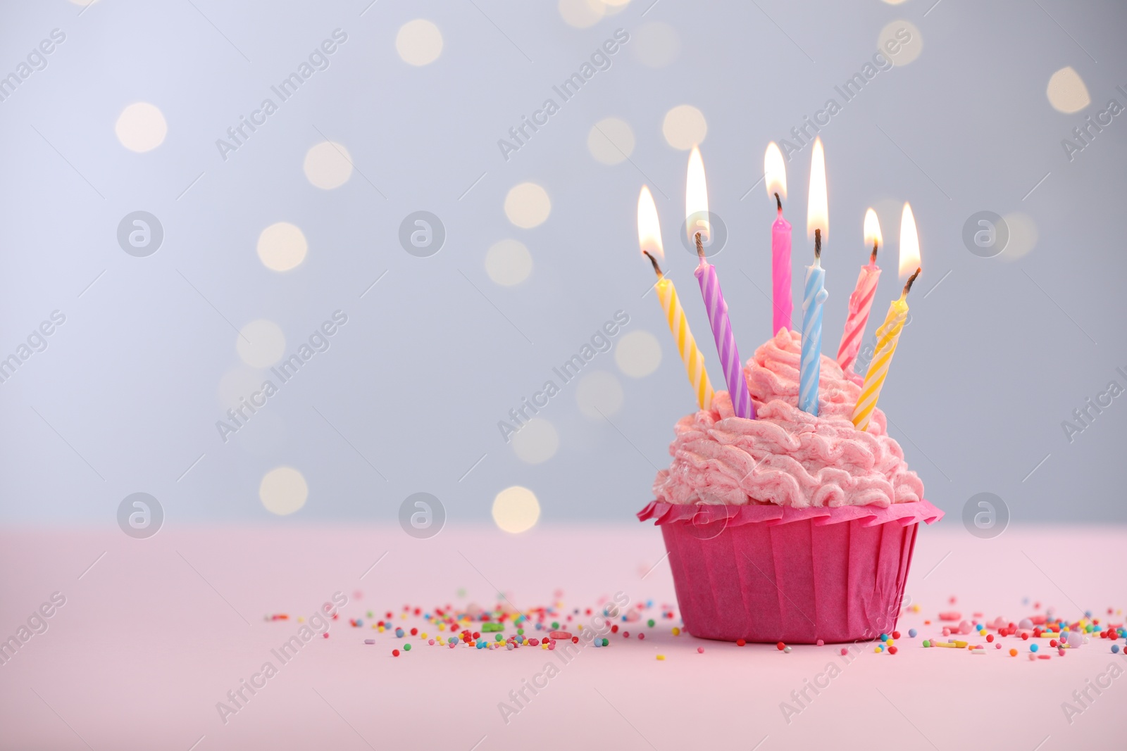 Photo of Burning candles in cupcake and sprinkles on table against blurred lights. Space for text