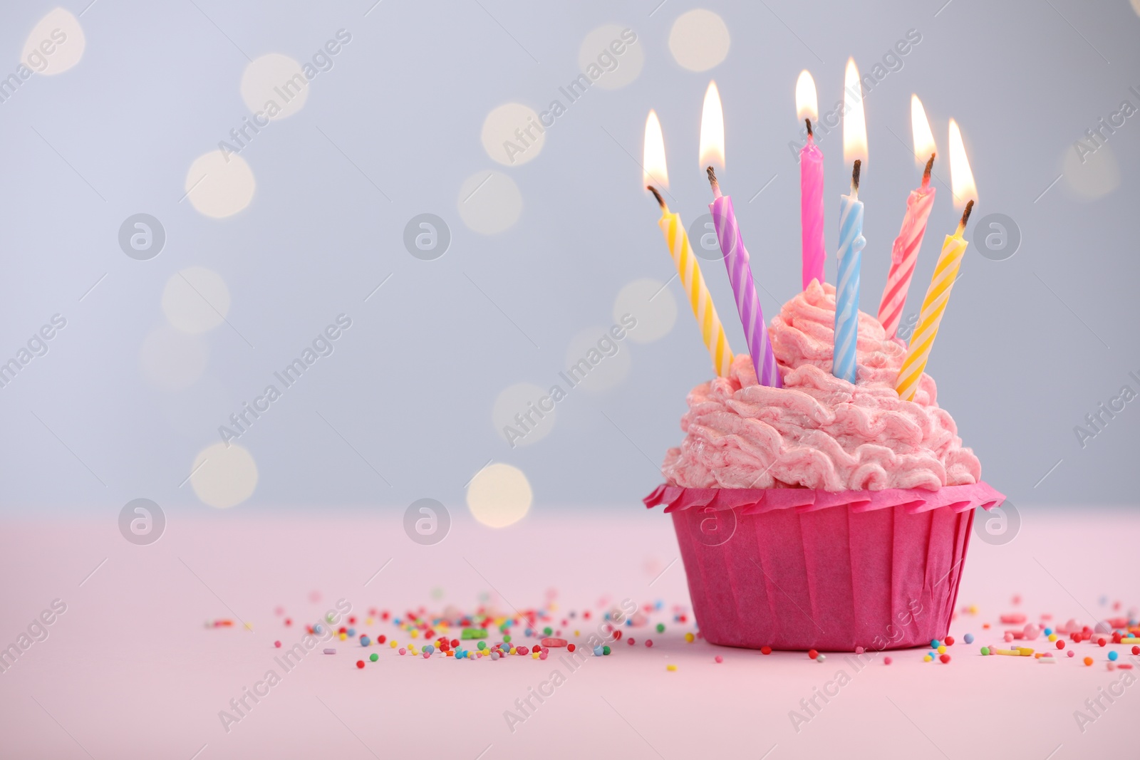 Photo of Burning candles in cupcake and sprinkles on table against blurred lights. Space for text