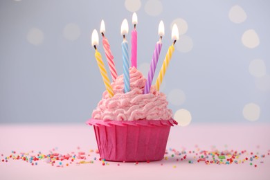Photo of Burning candles in cupcake and sprinkles on table against blurred lights