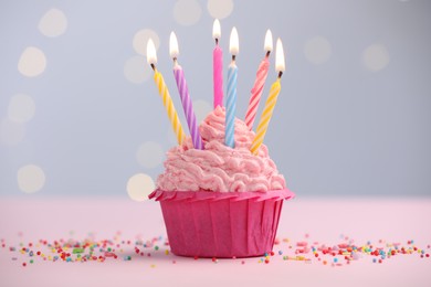 Photo of Burning candles in cupcake and sprinkles on table against blurred lights