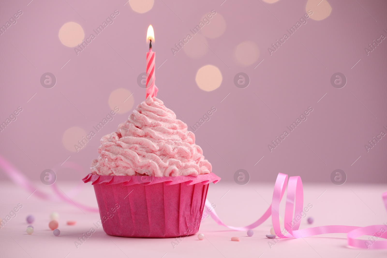 Photo of Burning candle in cupcake on table against pink background with blurred lights. Space for text