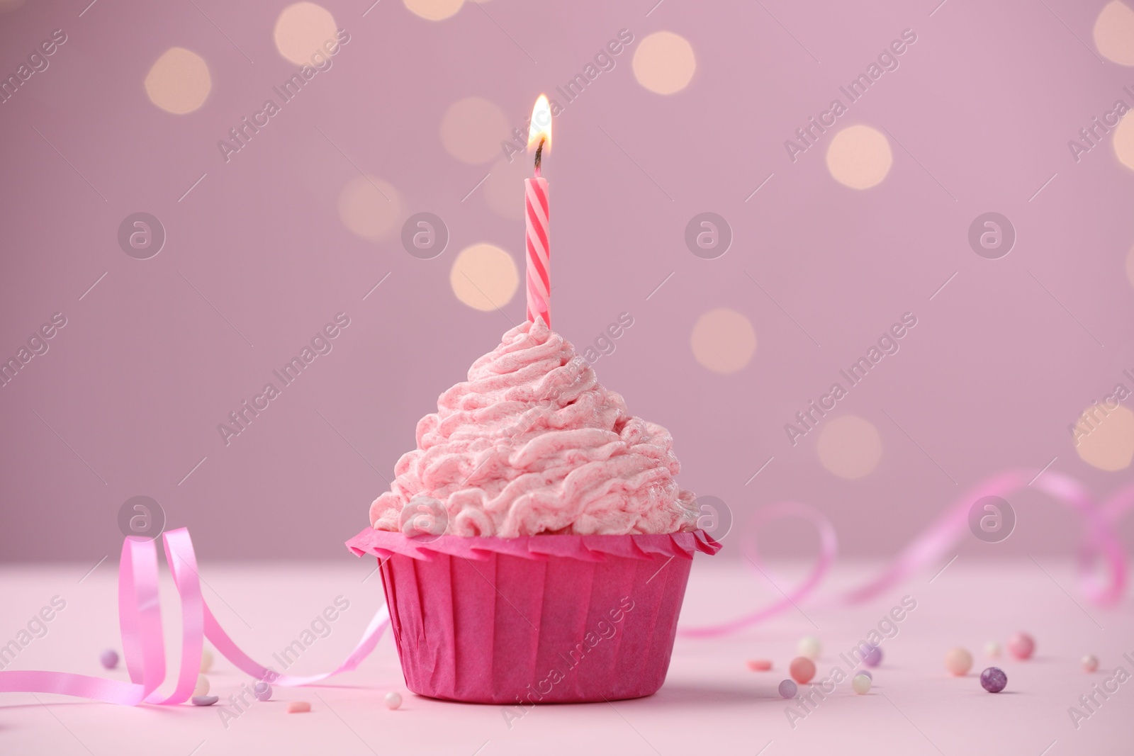 Photo of Burning candle in cupcake on table against pink background with blurred lights