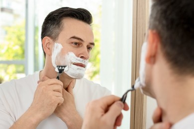 Photo of Handsome man shaving with razor near mirror at home