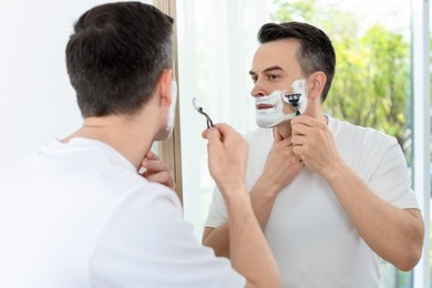 Photo of Handsome man shaving with razor near mirror at home