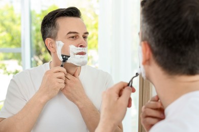 Photo of Handsome man shaving with razor near mirror at home