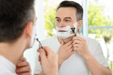 Photo of Handsome man shaving with razor near mirror at home