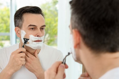 Photo of Handsome man shaving with razor near mirror at home