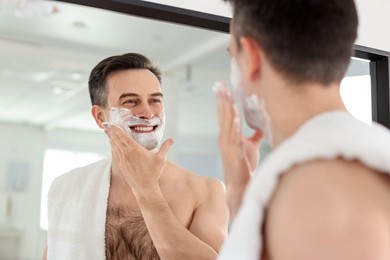 Photo of Smiling man applying shaving foam near mirror at home, low angle view