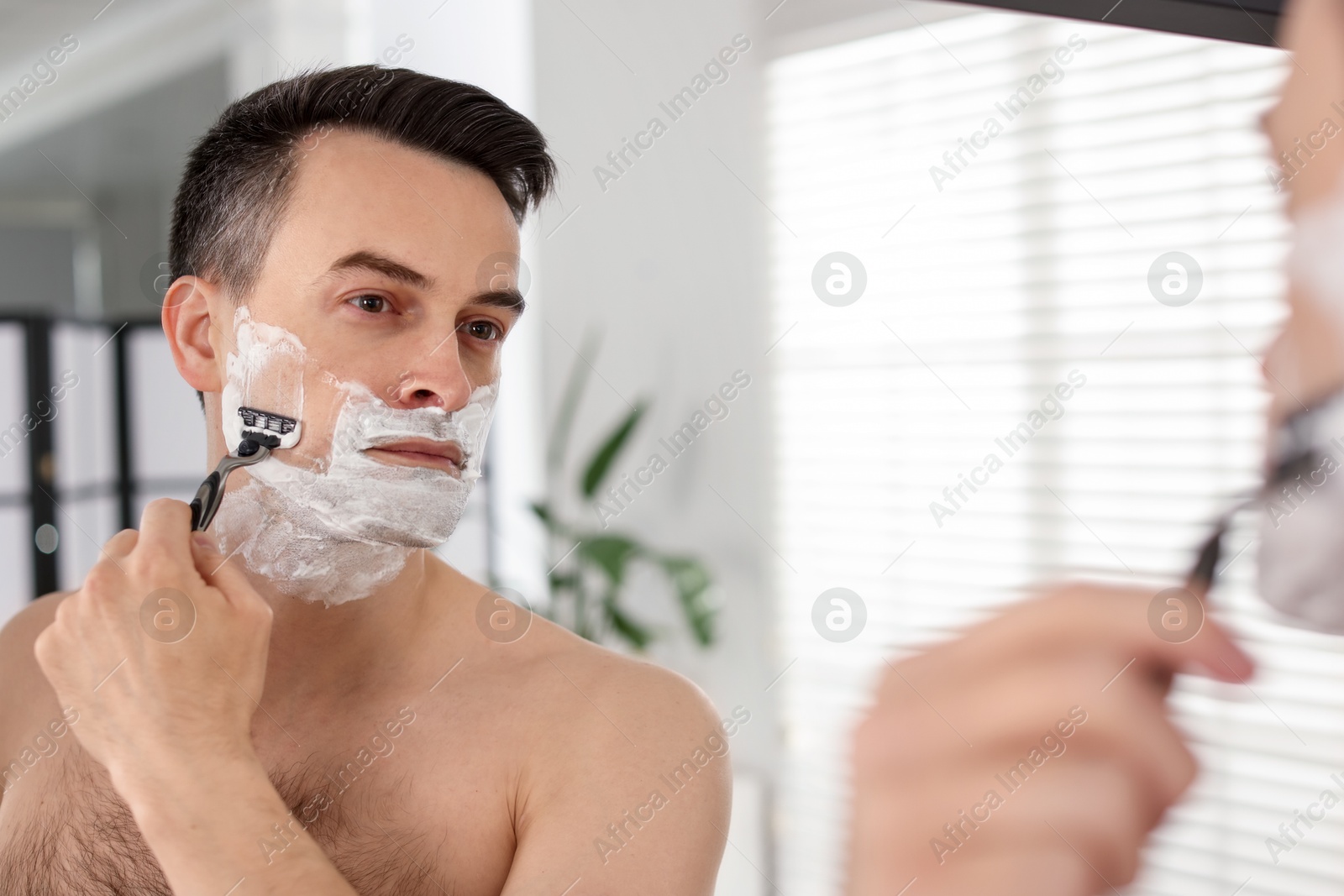 Handsome man shaving with razor near mirror at home Photo of Handsome man shaving with razor near mirror at home