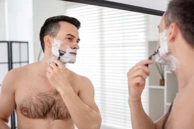 Photo of Handsome man shaving with razor near mirror at home