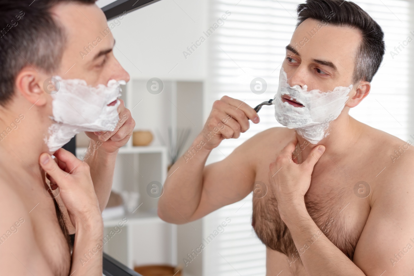 Photo of Handsome man shaving with razor near mirror at home