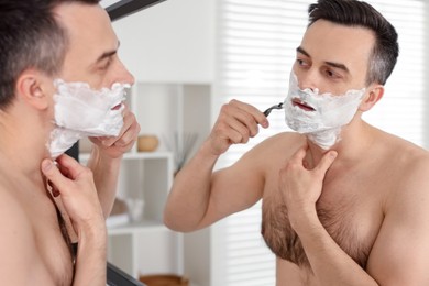 Photo of Handsome man shaving with razor near mirror at home