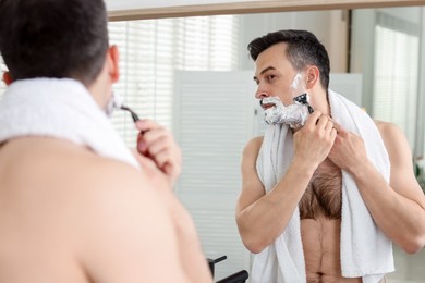Photo of Handsome man shaving with razor near mirror at home
