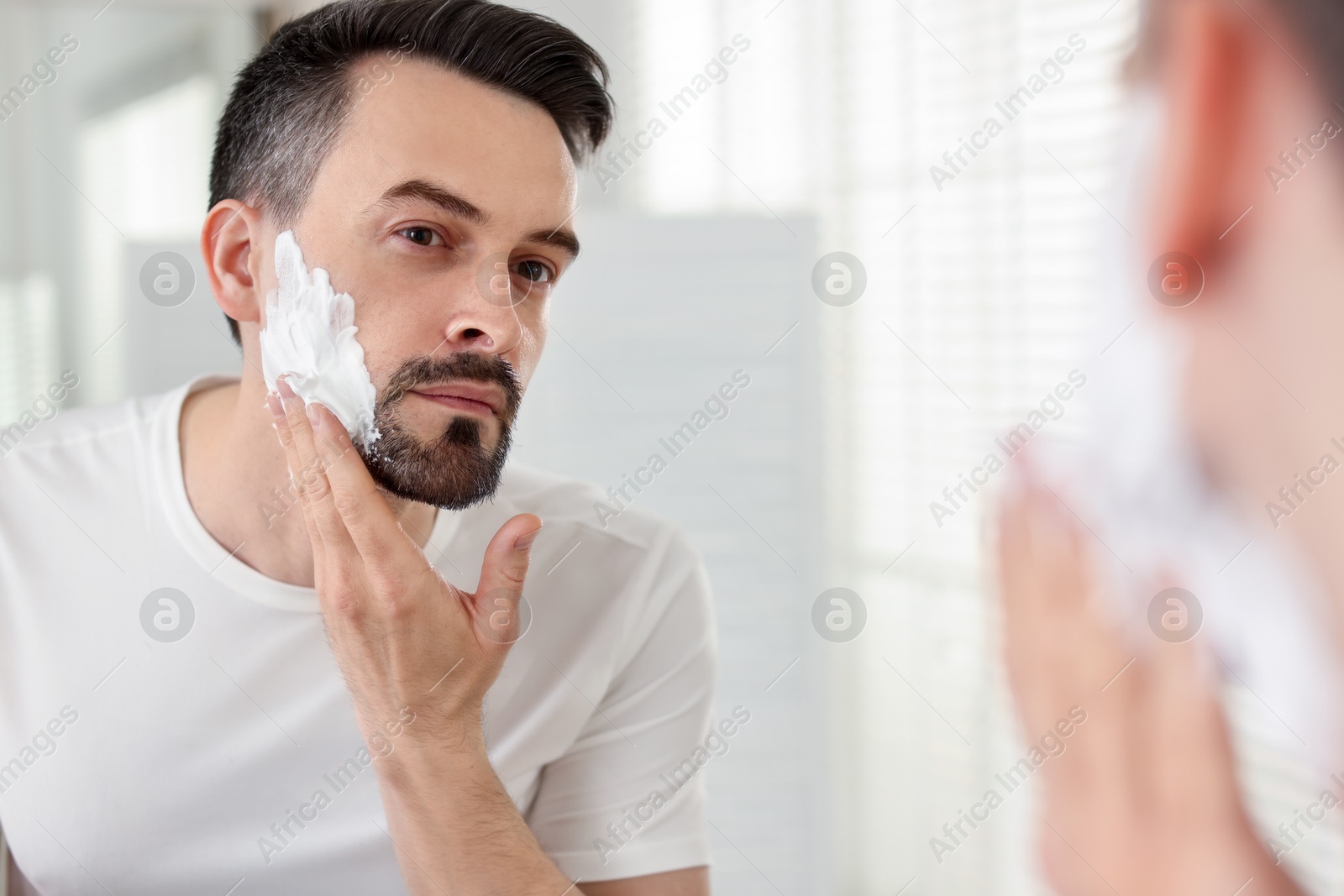 Handsome man applying shaving foam near mirror in bathroom Photo of Handsome man applying shaving foam near mirror in bathroom
