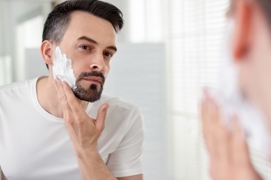 Photo of Handsome man applying shaving foam near mirror in bathroom