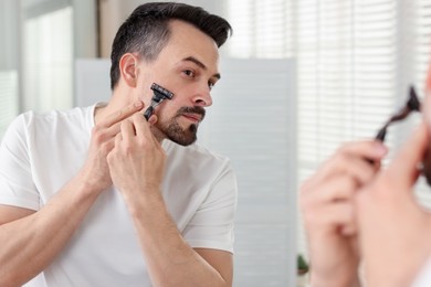 Photo of Handsome man shaving with razor near mirror at home