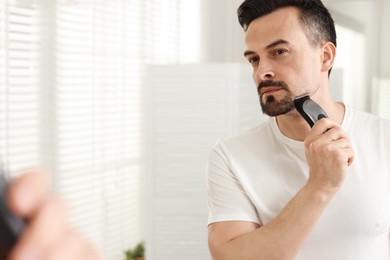 Photo of Handsome man shaving with trimmer near mirror at home. Space for text