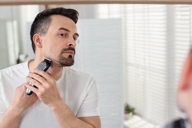 Photo of Handsome man shaving with trimmer near mirror at home. Space for text