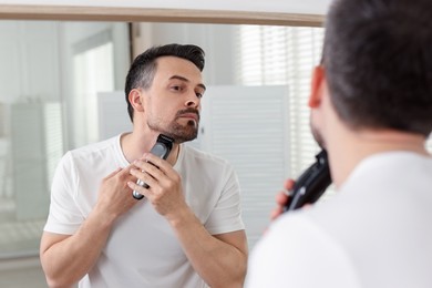 Photo of Handsome man shaving with trimmer near mirror at home