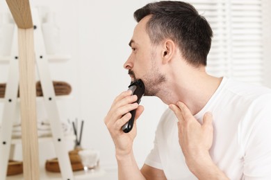 Photo of Handsome man shaving with trimmer near mirror at home