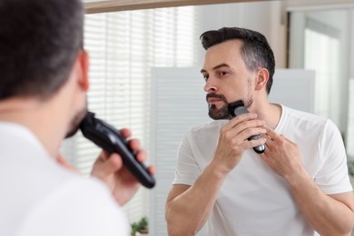 Photo of Handsome man shaving with trimmer near mirror at home