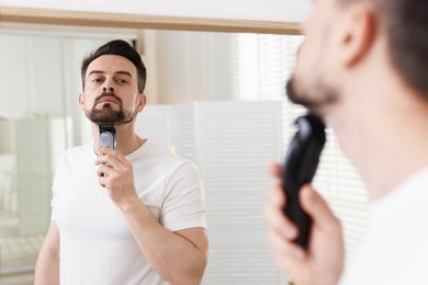 Photo of Handsome man shaving with trimmer near mirror at home