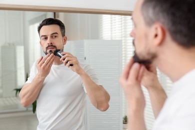 Photo of Handsome man shaving with trimmer near mirror at home
