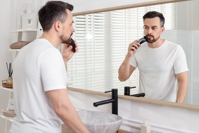 Photo of Handsome man shaving with trimmer near mirror at home