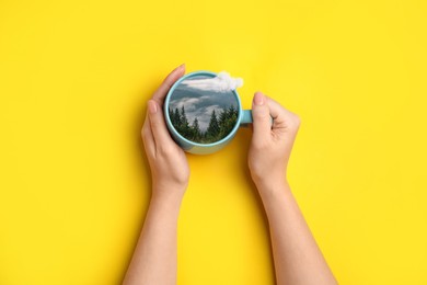 Image of Woman holding cup with mountain landscape inside on yellow background, top view. Cloud flying out of cup