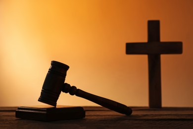 Photo of Silhouette of judge's gavel and cross on wooden table against pale orange gradient background, closeup