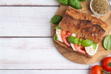 Photo of Tasty sandwich with mozzarella cheese, tomatoes and basil on white wooden table, flat lay. Space for text