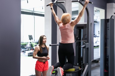 Photo of Senior woman training on power tower supervised by professional trainer in gym