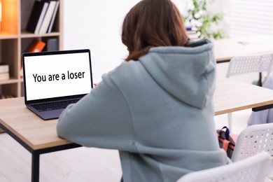 Photo of Cyber bullying. Upset teenage girl and laptop with text You Loser at desk indoors