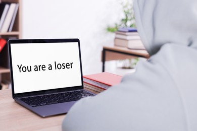Photo of Cyber bullying. Upset teenage girl and laptop with text You Loser at desk indoors, closeup