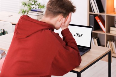 Photo of Cyber bullying. Upset teenage boy and laptop with text You Loser at desk indoors